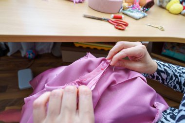 Dikiş etkinliği, ellerin ustalıkla pembe kumaştan bir elbise diktiği rahat bir çalışma alanında gerçekleşir. Makas ve iplikler yakınlarda, bir zanaat ortamı sergiliyorlar..