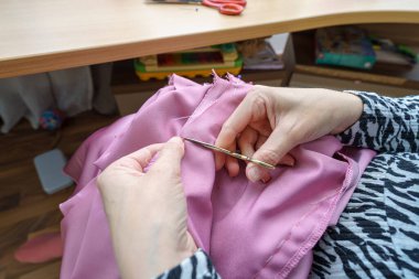 Bir insan, tahta bir masada otururken iplikleri kesmek için makas kullanarak kumaş dikmeye çalışıyor. Parlak doğal ışık odayı sarıyor, canlı pembe maddeyi vurguluyor..