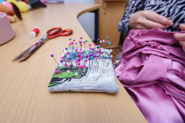 Renkli dikiş iğneleri bir yastığın üzerinde kümelenirken, bir insan evde dikiş düzeneğinde makas ile kumaş üzerinde çalışıyor. Atmosfer odaklanmış ve yaratıcıdır, bir zanaat faaliyetini yansıtır..