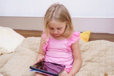 Çocuk, elindeki tablete odaklanmış, rahat bir yatakta oturuyor. Işıl ışıl aydınlatılmış oda sıcak bir atmosfer yaratıyor, boş zamanlarında konsantrasyonunu ve rahatlığını vurguluyor..