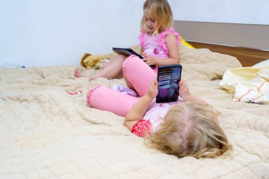 İki genç kız bir yatakta otururken cihazlarıyla meşgul oluyorlar. Büyük çocuk tabletine odaklanırken, küçük kız telefonuyla uzanmış, rahat bir atmosfer yaratıyor..