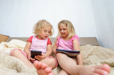 İki kız öğleden sonra bir yatakta oturup tabletlerine ve telefonlarına odaklanırlar. Güneş ışığı odayı dolduruyor, keyifli bir atmosfer yaratıyor ve birlikte keyifli bir anı paylaşıyorlar..