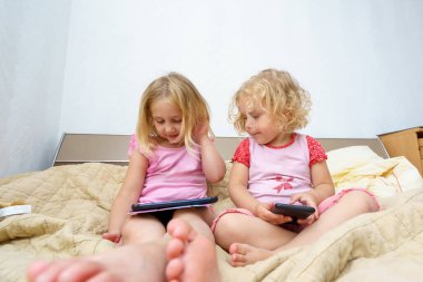 İki kız bir yatağa yakın otururlar, cihazlarına odaklanırlar. Parlak, rahat bir mekanda, ekranlarında yer alan bir bağlantı ve merak anlarını paylaşıyorlar..