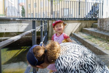 Şapka takan neşeli genç bir çocuk bir su kütlesinin yanında oynarken, yakınlardaki bir yetişkin başka bir göreve odaklanıyor. Güneş ışığı parlıyor, sıcak bir atmosfer yaratıyor..