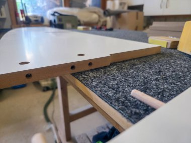 Beyaz bir panel, montaj için önceden delinmiş delikleri gösteren dokulu bir atölye masasının üzerinde duruyor. Ahşap işleme araçları arka planda görünür olup, gün boyunca etkin bir çalışma alanını gösterir.