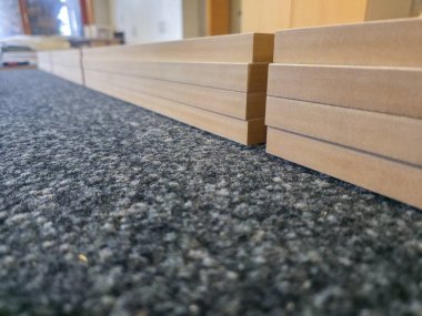 Bir atölyenin içindeki koyu renkli bir halıya düzgünce birkaç ahşap tahta dizilir. Ayarlar, malzemelerin sergilendiği bir inşaat veya zanaat projesinin hazırlanmasını öneriyor.