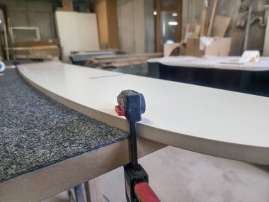 Becerikli bir zanaatkar düzgün bir bitiş elde etmek için kenarı zımparalarken kavisli bir ahşap paneli sabit tutmak için kıskaç kullanır. Çalışma alanı araçlar ve malzemelerle dolu..