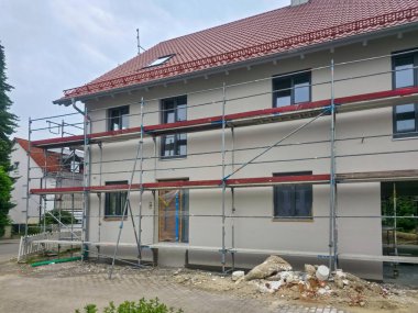 İşçiler, dış tarafında iskele bulunan bir konut binasını yeniliyorlar. Bulutlu hava, banliyö ortamına sessiz bir atmosfer ekliyor ve devam eden inşaatı vurguluyor..