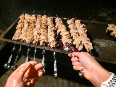 Yumuşak marine edilmiş et parçaları kızgın kömürlerin üzerinde ızgara yapıyor. Bir el, keyifli bir akşam barbeküsü sırasında yemek pişirme sürecini dikkatlice yönetiyor..