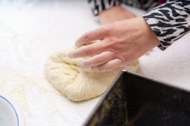 Bir insan sıcak bir mutfakta, un kaplı bir tezgahta hamur yoğurmak için özenle çalışıyor. Pürüzsüz hale geldiğinde hamur ellere ve dokusuna odaklanır..