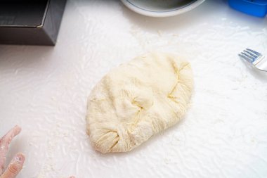 Hafif bir yüzeyde, un kaplı ellerle yoğurulduktan sonra bir hamur yığını dinlenir. Yakınlarda bir çatal var, pişirme sürecini işaret ediyor. Bu ev mutfağı sahnesi sıcaklık ve geleneği çağrıştırıyor..