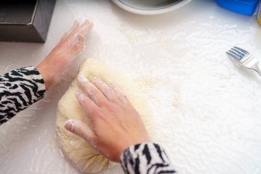 Elleri un kaplı, bir hamur topunu pürüzsüz bir tezgahta yoğurur. Mutfak gereçleri ve kâseleri görülebilir durumda, bu da gündüz yemek hazırlığını gösteriyor..