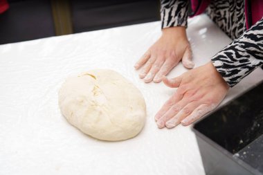 Birisi bir ev mutfağında temiz beyaz bir yüzeye hamur topu yoğuruyor. Kişi şık, desenli bir bluz giyer ve pişirmeye hazırlanmaya odaklanır..