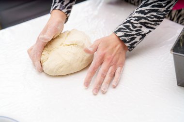 Elleri sıcak bir ev mutfağında beyaz bir tezgahta ekmek hamuru yoğuruyor. Un tozu mevcut, bu da devam etmekte olan bir pişirme oturumunu gösteriyor.