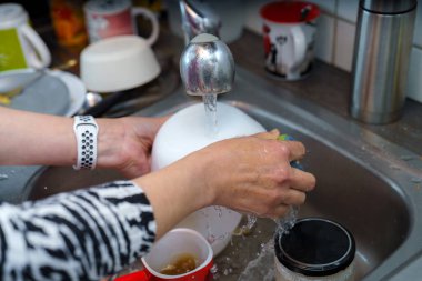 Eller mutfak lavabosunda akan suyun altında büyük beyaz bir kase ovalıyor. Yakınlarda, çeşitli bardaklar ve çatal bıçaklar etrafa saçılmış durumda, bu da yoğun bir yemek pişirme seansına işaret ediyor..
