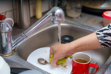 Bir el mutfak lavabosunda akan suyun altındaki tabağı ovalıyor. Yakınlarda, kırmızı bir fincan çay içerken, tezgahta birkaç kap görülebilir. Bu günlük angaryada temizlik vurgulanıyor.