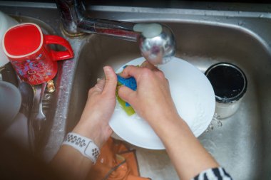 Elleri mutfak lavabosunda akan suyun altında süngerle tabağı ovalıyor. Parlak renkli bardaklar ve konteynırlar lavabonun etrafını sarıp, canlı bir iç mekan yaratıyorlar..