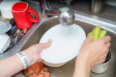 Eller bulaşık dolu bir mutfak lavabosunda akan suyun altında beyaz bir tabağı ovalıyor. Ev işlerinin hafta sonu bittiği bir evde rahat bir atmosfer görülür..