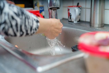 Bir insan mutfak lavabosunda bulaşık yıkıyor, sünger sıkmak için elini kullanıyor. Musluktan su akıyor, çeşitli mutfak eşyalarıyla çevrili paslanmaz çelik havuza sıçrıyor..
