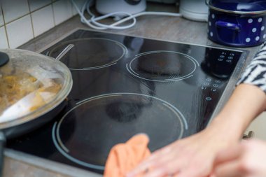 Bir insan yemek hazırladıktan sonra bir ev mutfağında sobayı temizliyor. Mutfakta mutfak gereçleri ve mutfak gereçleri sergileniyor, yoğun bir yemek ortamı vurgulanıyor.