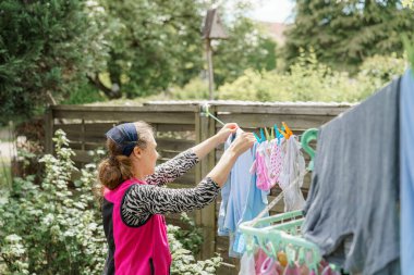 Bir kadın bahçesindeki çamaşır ipine yeni yıkanmış giysileri dikkatlice asar. Parlak güneş, kıyafetlerin rengini ve çevreleyen yeşilliği güçlendirir ve sakin bir atmosfer yaratır..