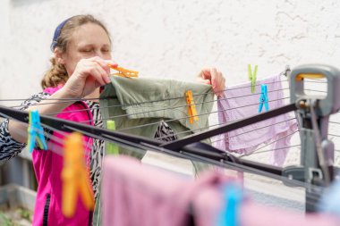 Saç bandı takan bir kadın, arka bahçesindeki çamaşır ipine yeni yıkanmış giysiler asıyor. Güneş etrafını aydınlatıyor, neşeli bir atmosfer yaratıyor..