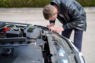 Genç bir adam, motor parçalarını incelemek için arabasının kaputunun üzerine eğiliyor. Açık bir öğleden sonra ve o bir sorunu teşhis etmek için özenle çalışırken güneş aracın üzerinde parlıyor..