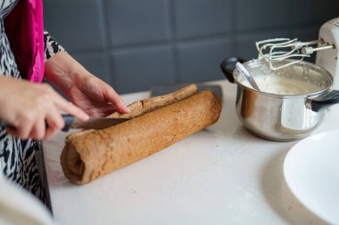 Bir fırıncı, sıcak bir mutfakta ustalıkla çikolatalı pasta kesiyor. Yanında da pişirme aletleri ve bir kase krema var..
