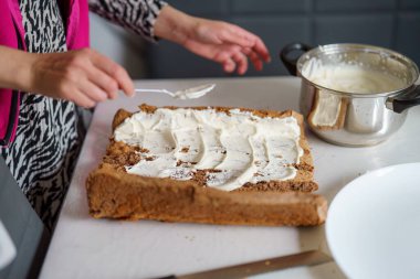 Parlak bir mutfakta, bir insan taze pişmiş bir tatlının üzerine beyaz bir tezgahta krema sürer. Atmosfer sıcak ve davetkar. Ev yapımı ikramlara olan sevgisini gösteriyor..
