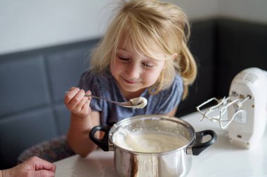 Genç bir kız, mutfak masasında otururken metal bir kâsede krema karıştırma işleminden zevk alıyor. Bir kaşık tutuyor ve gülümsüyor, mutfak malzemeleriyle çevrili..