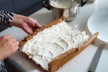 Bir insan, tezgahtaki taze pişmiş zencefilli kurabiye tabanına pürüzsüz bir krema katmanı sürüyor. Parlak mutfakta tencere ve aletler var..