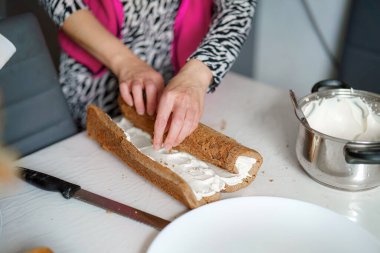 Bir insan şık bir mutfakta kremayla dolu bir pastayı ustalıkla sarıyor. Parlak tezgahta pişirme araçları ve malzemeler yer alıyor..