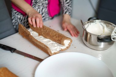 Bir insan pasta katmanına krema dökerek çikolatalı pasta hazırlar. Dekor rahat bir mutfak, içinde krem şanti var..