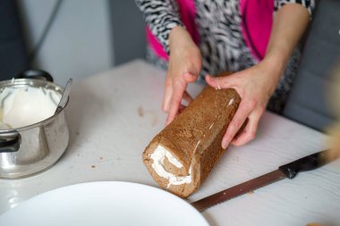 Parlak bir mutfakta kremayla dolu çikolatalı keki ustalıkla saran bir insan. Eller, mükemmel bir yuvarlanma yaratmak için nazikçe baskı uyguluyor..