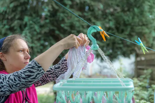 Bir kadın bahçesindeki kurutma askısına yeni yıkanmış giysileri dikkatlice asar. Parlak güneş ışığı, kumaşın canlı renklerini gösteren sahneyi aydınlatır..