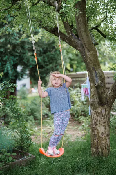 Küçük bir çocuk arka bahçedeki bir ağaçtan sarkan salıncakta neşeyle oynuyor. Güneş parlıyor, yeşillikler arasında sıcak bir atmosfer yaratıyor..