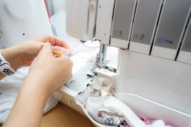Eller kumaşı dikiş makinesinde ustalıkla yönlendirir. Etrafında iplikler ve kırıntılar vardır. Çalışma alanı iyi ışıklandırılmış ve giysi yapımında kullanılan araçları gösteriyor.