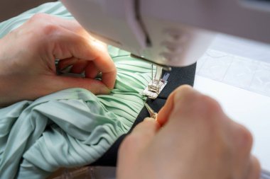 Eller dikiş makinesi iğnesinin altındaki soluk yeşil pileli kumaşı ustalıkla manipüle ediyor. Çalışma alanı düzenlenmiştir ve giysi yapımının karmaşık sürecini vurgulamaktadır..