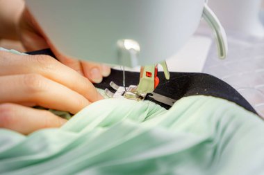 Eller dikiş makinesi iğnesinin altında kumaşa ustalıkla rehberlik eder. Gündüz saatlerinde iyi aydınlatılmış bir stüdyoda giysi yapma sanatını vurgular. Hassas dikiş teknikleri gösterir..