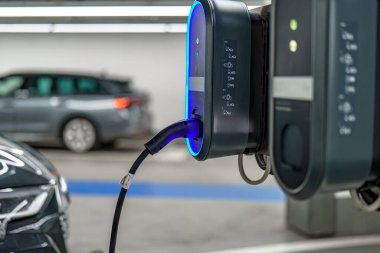 Şık bir elektrikli araç geniş bir otoparkta bulunan bir şarj istasyonuna bağlanıyor. İstasyonda parlak mavi bir aydınlatma var..