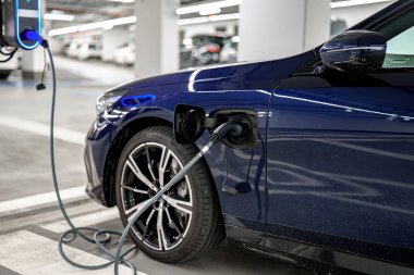 Parlak mavi bir elektrikli araç, iyi aydınlatılmış bir otoparkta şarj istasyonuna bağlanıyor. Kurulumda parlak arabalar ve temiz hatlar, çevre dostu teknoloji sergileniyor.