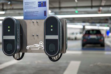 İyi aydınlatılmış bir yeraltı otoparkındaki metal bir panele iki elektrikli araç şarj ediliyor. Arka planda şarj altyapısını vurgulayan elektrikli bir araba görülebilir..