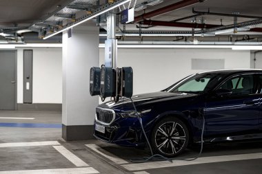 Şık bir elektrikli araç, iyi aydınlatılmış bir yeraltı garajındaki şarj istasyonlarının yanına park edilmiş. Modern altyapı çevre dostu sürücüler için kolaylık sunuyor.