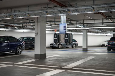 Elektrikli araçlar için bir şarj istasyonu iyi aydınlatılmış bir yeraltı otoparkında belirgin bir şekilde sergilenmektedir. Çevredeki arabalar düzgün park edilmiş, modern tasarım ve işlevsellik sergileniyor..