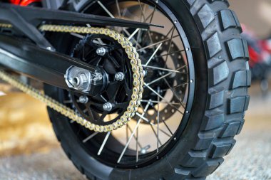 Bir motosikletin arka tekerlek ve zincirinin atölye içindeki yakın görüntüsü. Odak noktası karmaşık mekanikler, lastik izi ve metal bileşenleri keskin detaylarıyla gösteriyor..