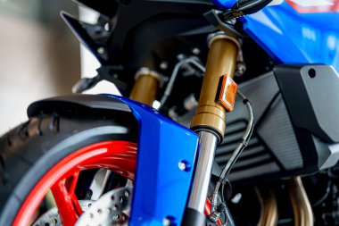 Yakından bakıldığında altın sarısı askılı çatallar, kırmızı tekerlekler ve mavi motor kaportası görülüyor. Dekor, hareketli bir şehir ortamında modern tasarım ve mühendislik mükemmelliğini yansıtıyor..