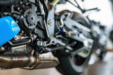 Tamirciler, iyi aydınlatılmış bir garajda motora ve egzoz bileşenlerine odaklanan bir motosiklet üzerinde çalışıyorlar. Araçlar ve parçalar düzenlendi, bakım görevleri ve güncellemeler için hazırlar.