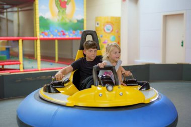 İki çocuk, kapalı bir eğlence merkezinde sarı bir çarpışan arabaya binerken gülümsüyor. Renkli ortam gün ortasında neşeli atmosferi artırır..