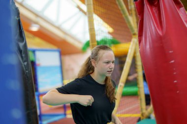 Bir kız fiziksel antrenman yapıyor, canlı bir kapalı oyun alanında spor çantasına yumruklar atıyor. Oyuncak yapılar ve aktivitelerle dolu..