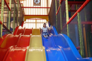 Bir kız, öğleden sonra ışığında canlı kırmızı, sarı ve mavi slaytlarda kayarak kapalı bir oyun alanında zaman geçirmekten zevk alır. Atmosfer canlı ve neşeli, oynamak için mükemmel..